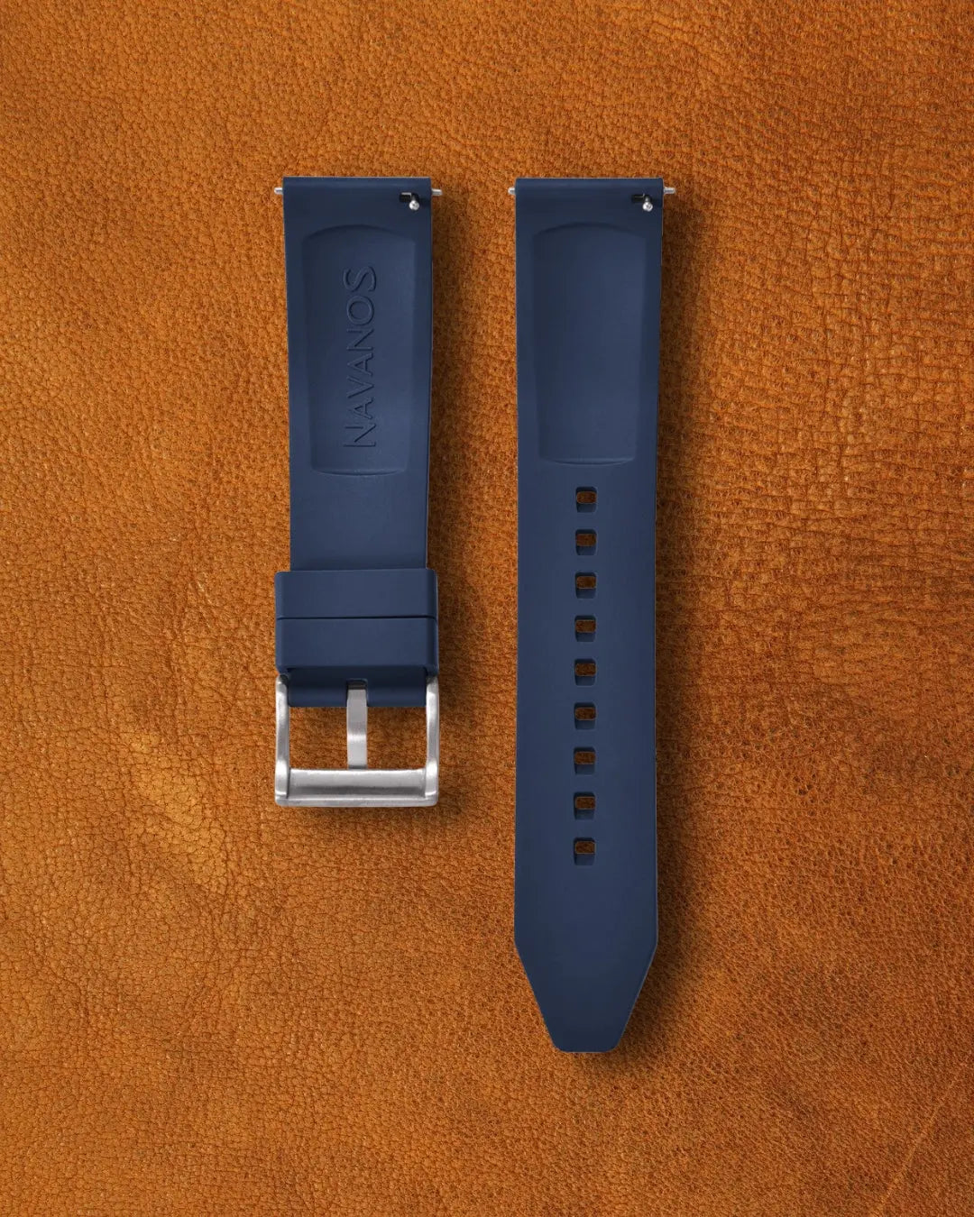 Nautical blue FKM rubber watch strap for Navanos watches with embossed "NAVANOS" branding, stainless steel tang buckle, quick-release spring bars, and multiple adjustment holes, displayed on brown suede background.