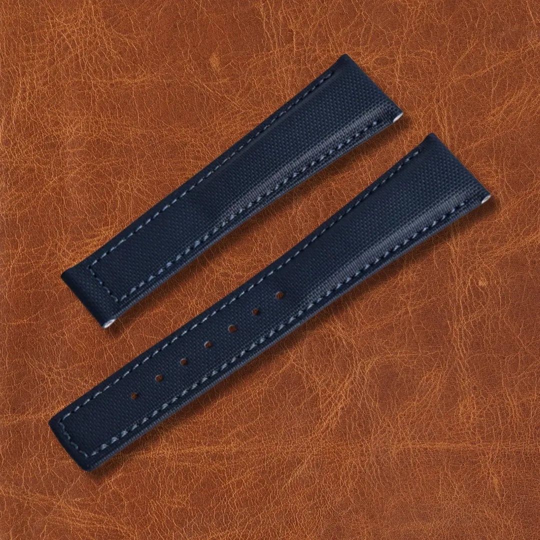 Nautical blue sailcloth watch strap with white stitching and adjustment holes, displayed on a brown leather background.