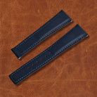 Nautical blue sailcloth watch strap with white stitching and adjustment holes, displayed on a brown leather background.