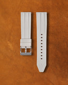 Coral white Ravaro Diver FKM rubber watch strap with ribbed design, embossed "Rayaro DIVER" keeper, trident logo on tip, stainless steel tang buckle engraved "NAVANOS", quick-release spring bars, and adjustment holes, displayed on brown suede background.
