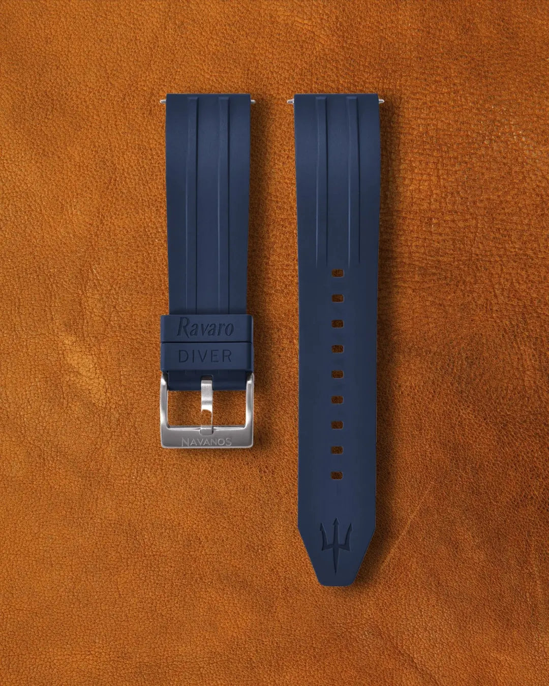 Nautical blue Ravaro Diver FKM rubber watch strap with embossed "Ravaro DIVER" keeper, trident logo on tip, stainless steel tang buckle engraved "NAVANOS", quick-release spring bars, and adjustment holes, displayed on brown suede background.