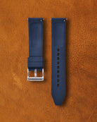 Nautical blue FKM rubber watch strap for Navanos watches with embossed "NAVANOS" branding, stainless steel tang buckle, quick-release spring bars, and multiple adjustment holes, displayed on brown suede background.